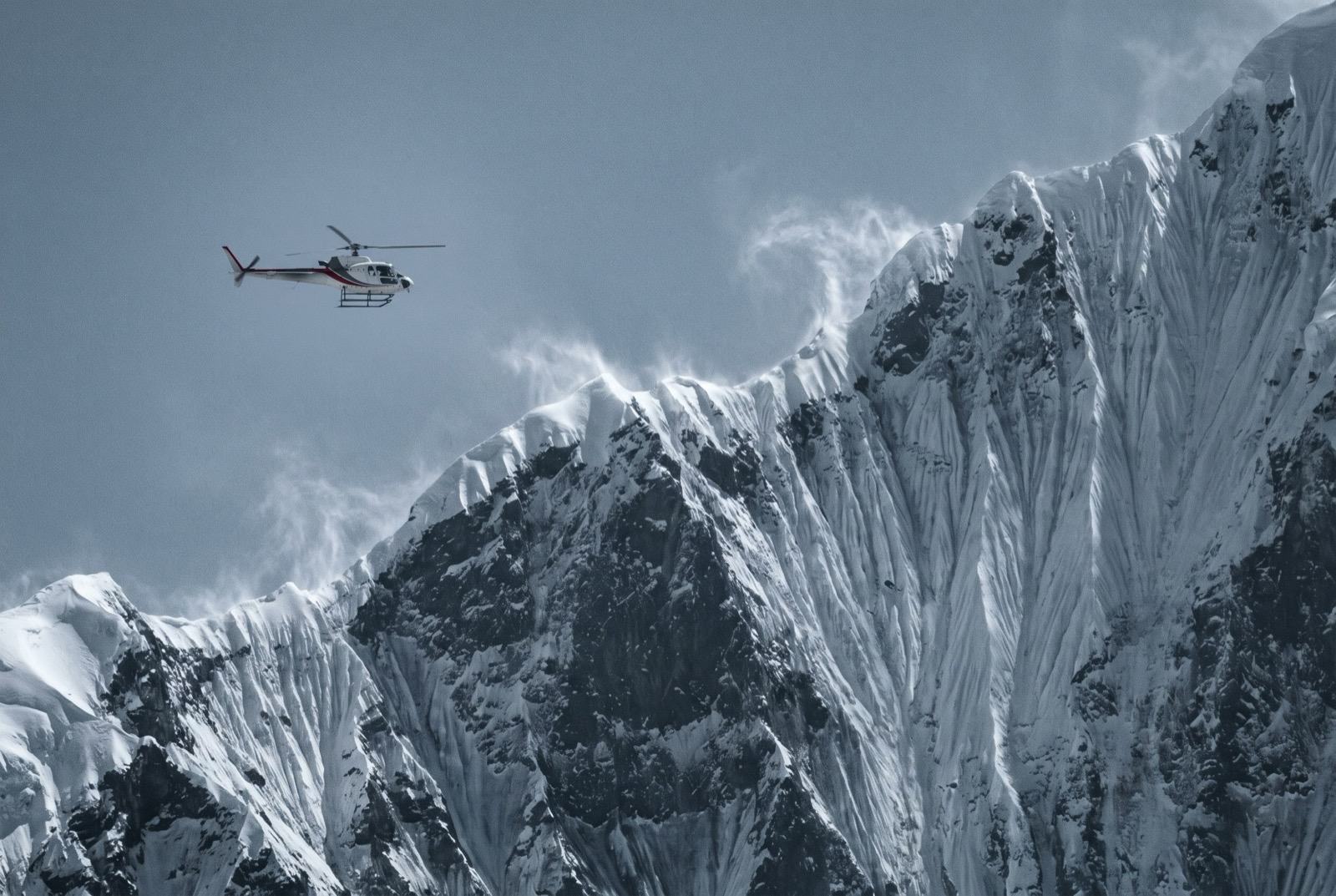 A single-engine helicopter, small in the frame, flying below an enormous wind-scoured face of snow and rock with spindrift streaming off the ridge above it.