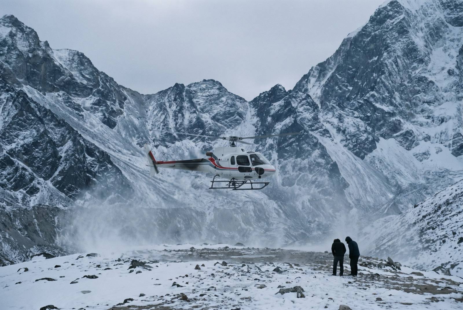 A single-engine helicopter lifting a few metres off a high landing zone, downwash driving snow and grit outwards, two figures standing back with their backs to the camera.