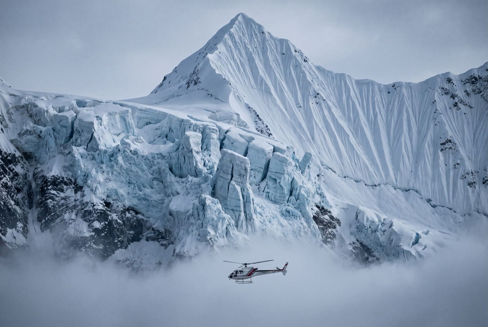 A small white helicopter crossing below an overhanging blue ice cliff on a sharply pointed fluted peak.