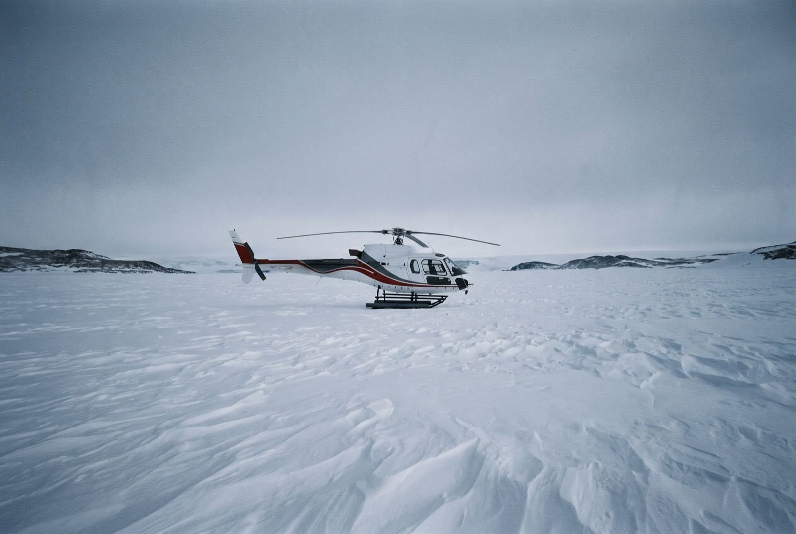 A white helicopter alone on an empty wind-scoured snow plain ringed by low dark ridges.