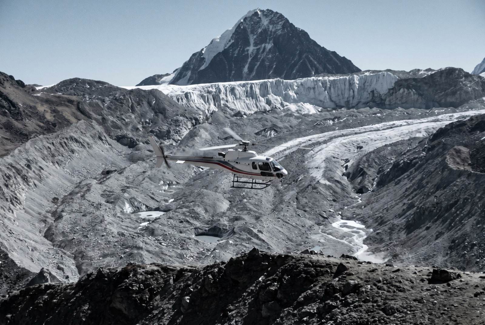 A white helicopter flying above a grey glacial moraine, a crevassed icefall and a dark summit pyramid behind it.