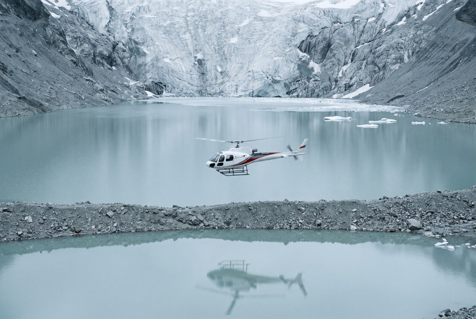 A white helicopter crossing above a still grey-turquoise glacial lake beneath an immense rock and ice wall.