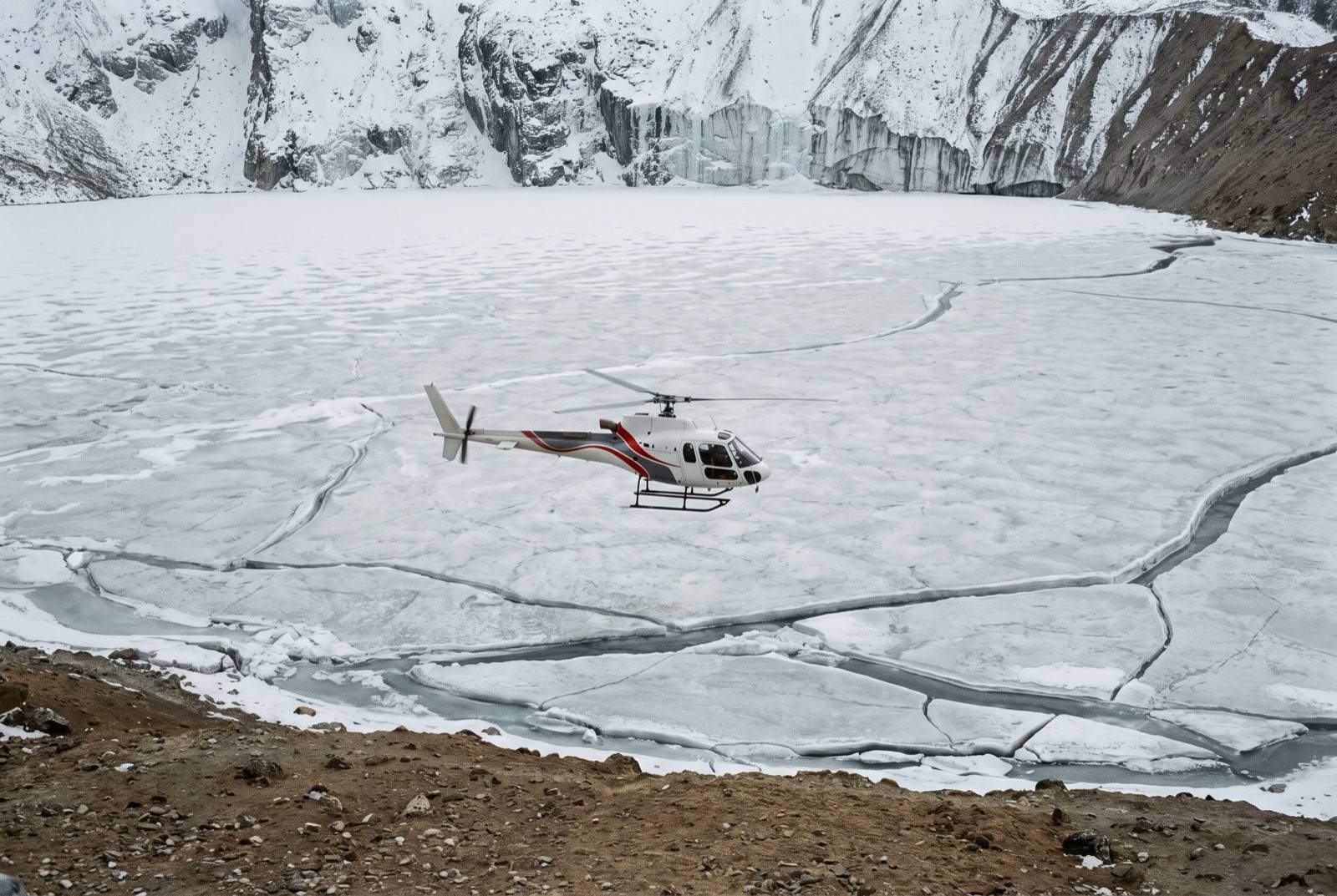 A white helicopter flying above a frozen high-altitude lake cracked into plates, a sheer ice-plastered wall beyond.