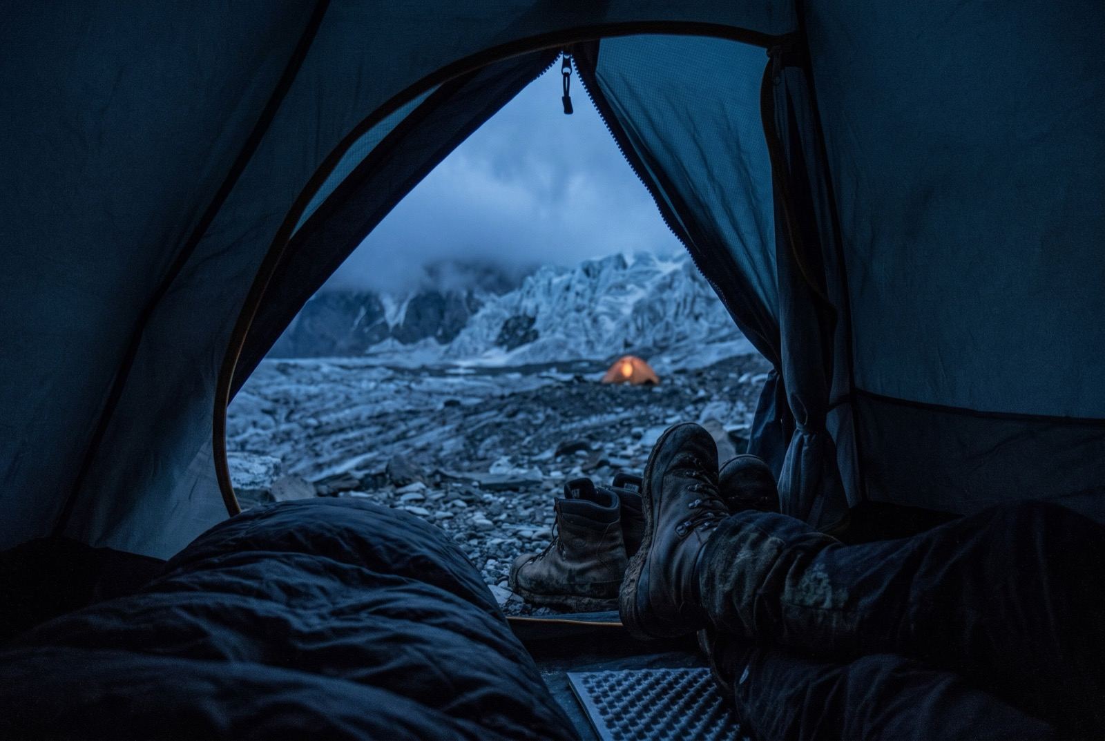 From inside a tent at dusk: a half-open zip, boots and a sleeping bag along the bottom edge, a glacier and one lit tent beyond.