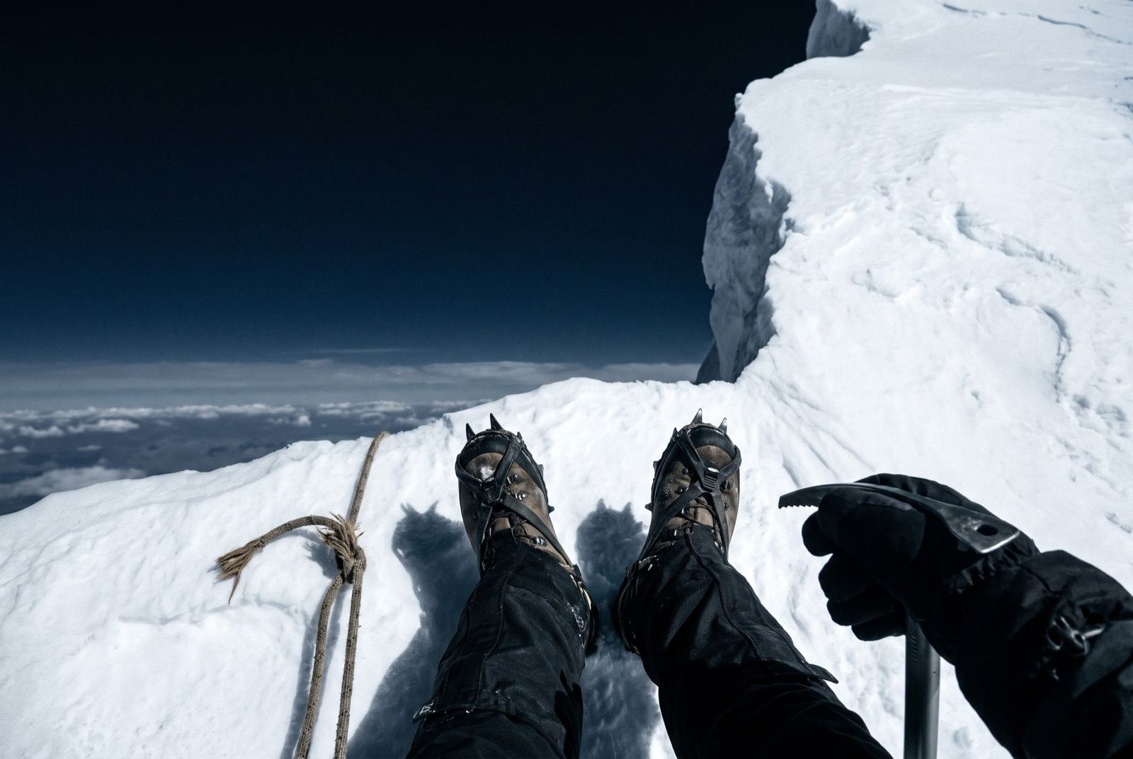 Looking down at crampons on a snow ridge where it stops, the frayed end of a rope alongside, dark sky and a cloud floor far below.