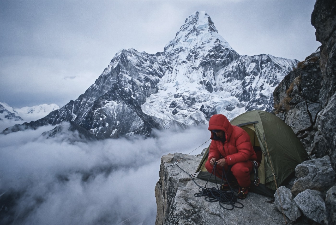 A tent pitched on a narrow ledge beneath Ama Dablam's summit spire and hanging glacier, a climber in a red jacket coiling rope at the door.