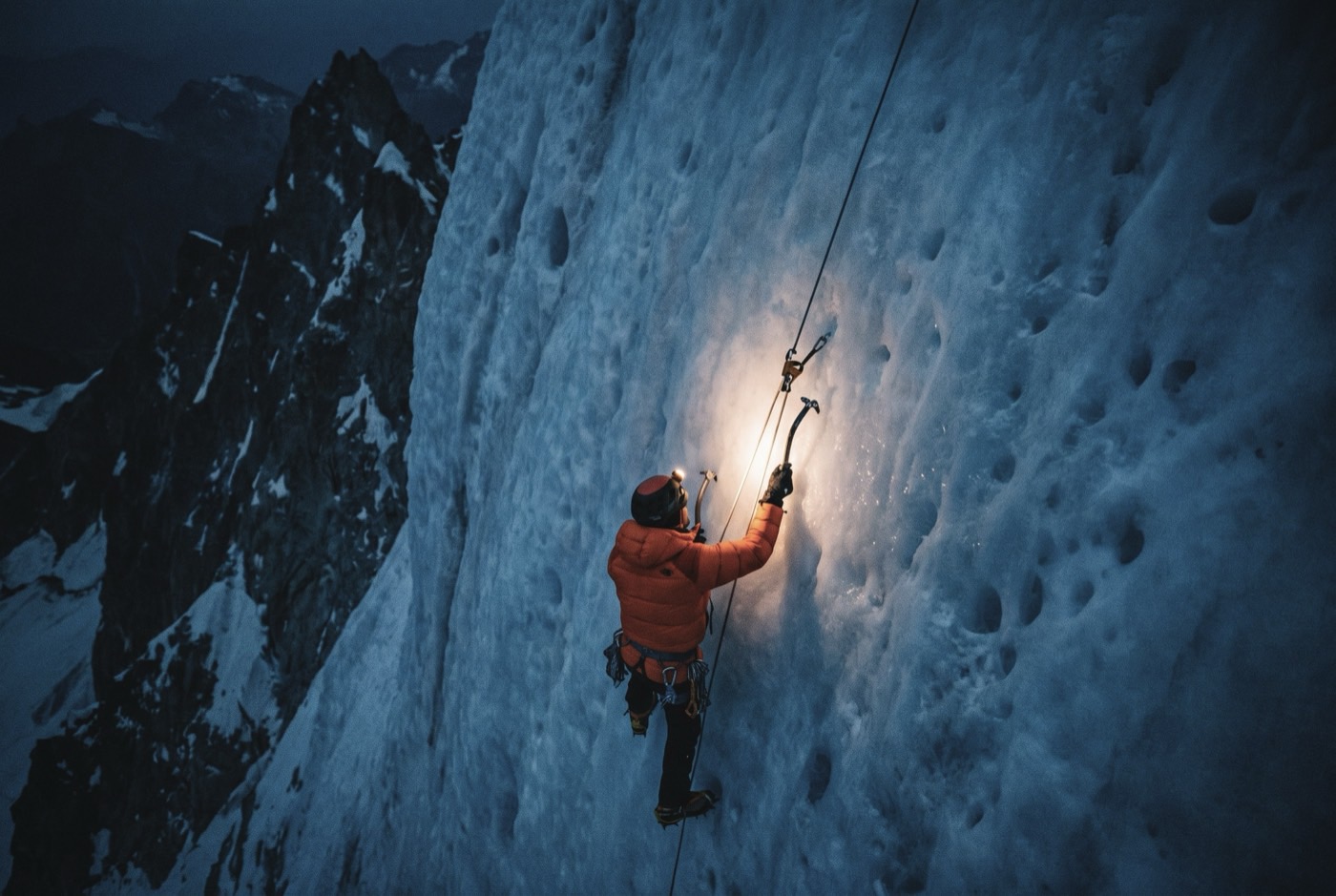 A climber in an orange down jacket ascending fixed ropes on a steep blue-ice headwall before dawn, lit only by their own head torch.