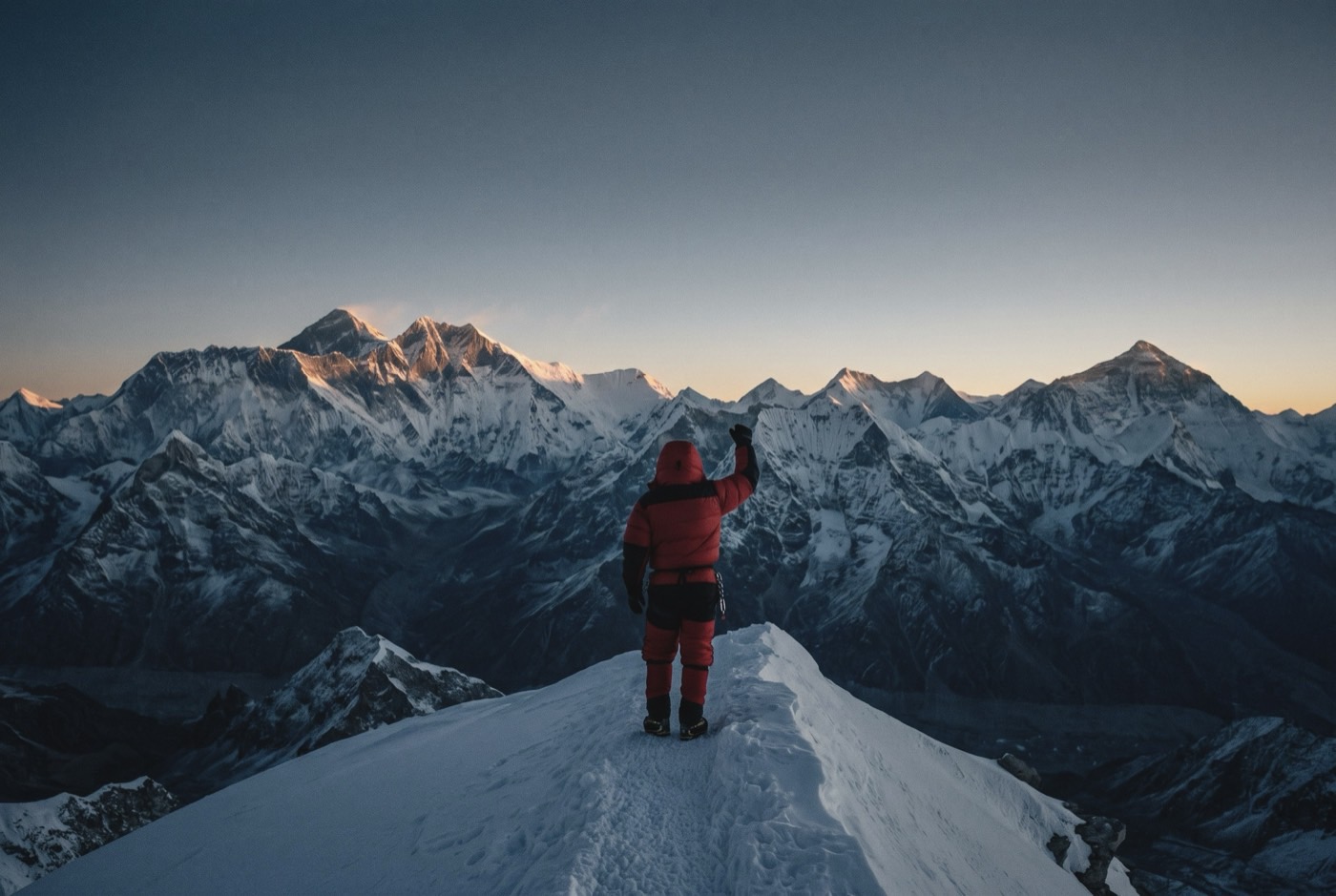 A lone climber standing on a wide snow summit ridge at dawn, tiny against a horizon of 8,000-metre Himalayan peaks.