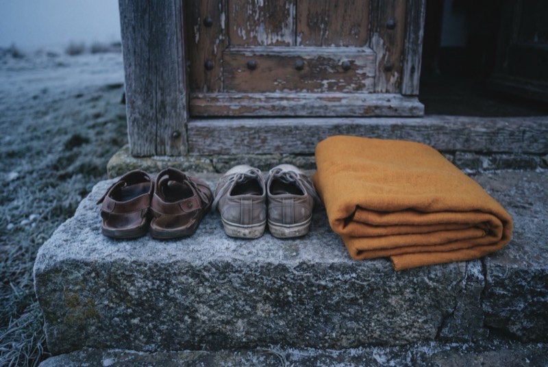 Worn leather sandals and canvas shoes left on a frosted stone step outside a closed wooden door, a folded saffron blanket beside them.