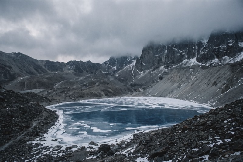 Tilicho Lake frozen in its bowl of dark shattered rock beneath cloud-covered walls